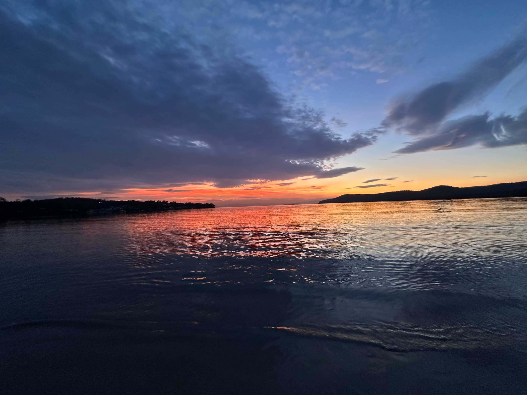 Sunset over the Tasman Peninsula — soft orange and blush light on still water, a horizon of low hills.